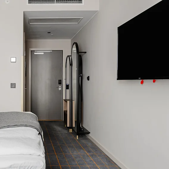 A neatly made bed with white linens and gray accent blanket, adjacent to a dark TV screen, in a modern, minimalist hotel room with abstract wall art and corridor view.