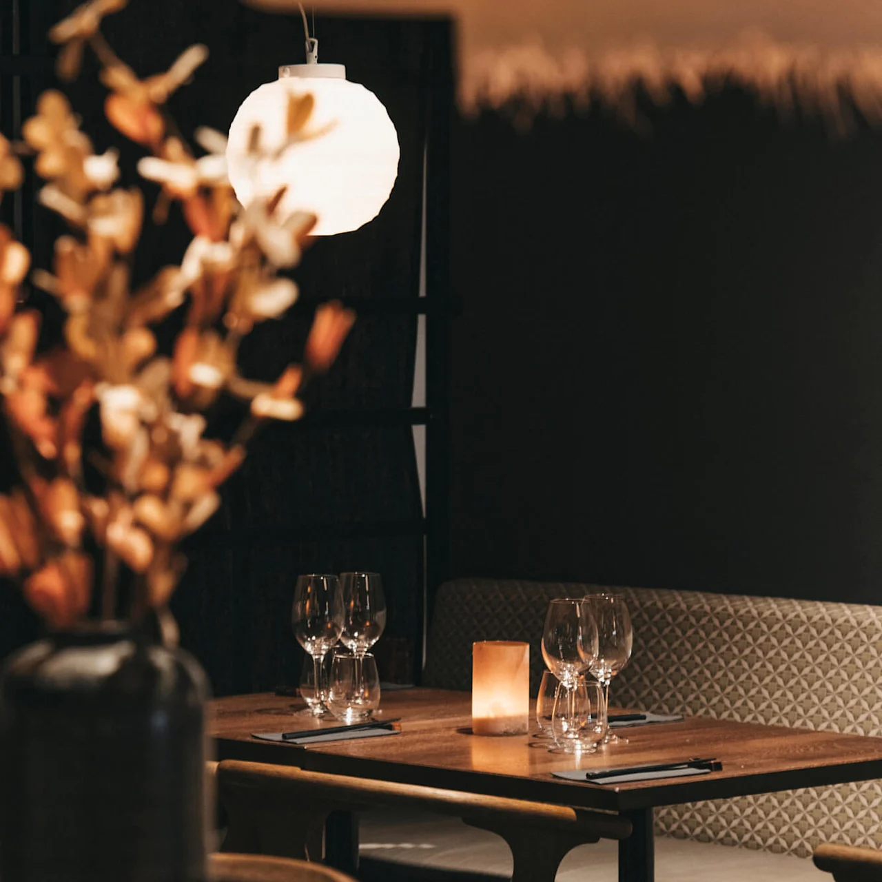 En intim restaurantinnstilling, med et varmt lys fra en hengende sfærisk lampe og stearinlys, som fremhever et tomt trebord med vinglass.
