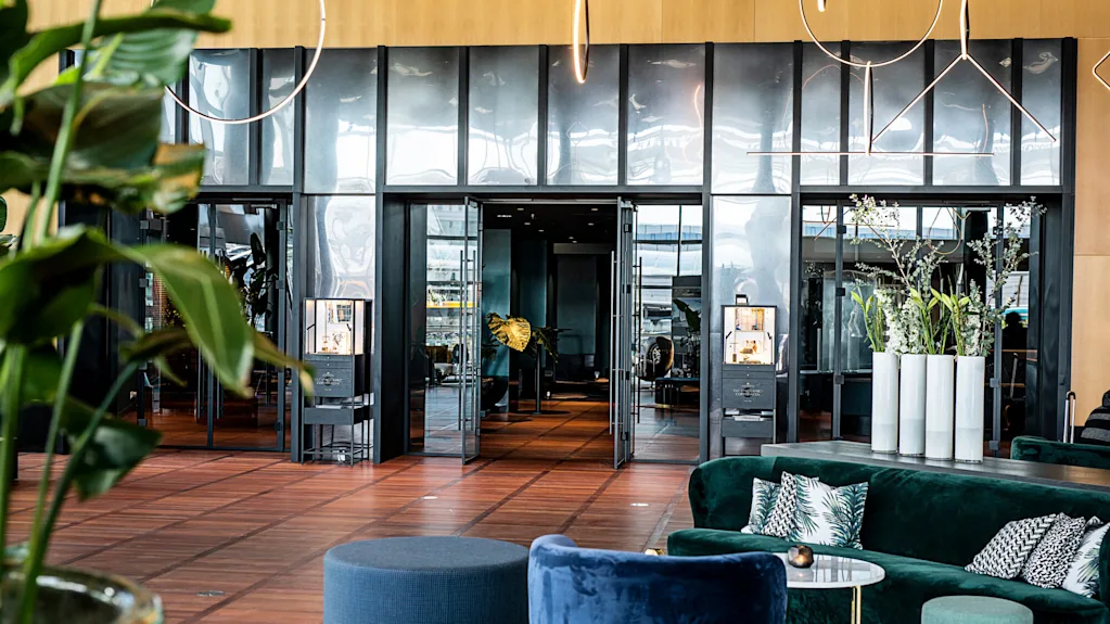 Luxurious hotel lobby featuring plush seating and elegant decor. Large glass doors open to a stylish interior with plants and ambient lighting, creating a sophisticated and welcoming atmosphere.