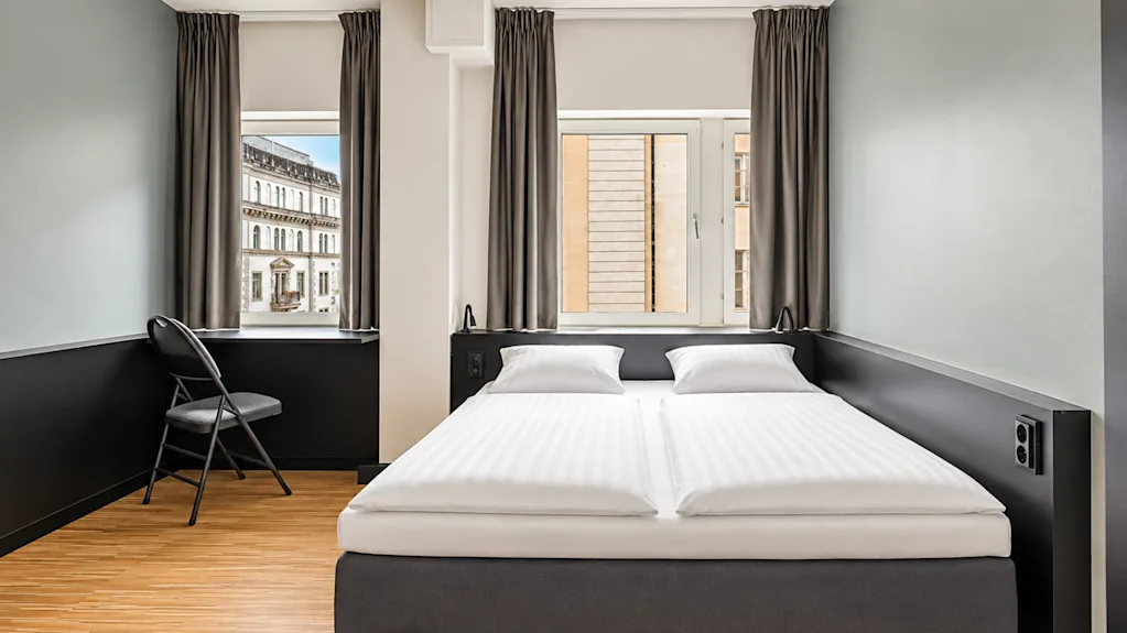 Modern hotel room featuring a double bed, light walls, wooden flooring, and two windows letting in daylight.