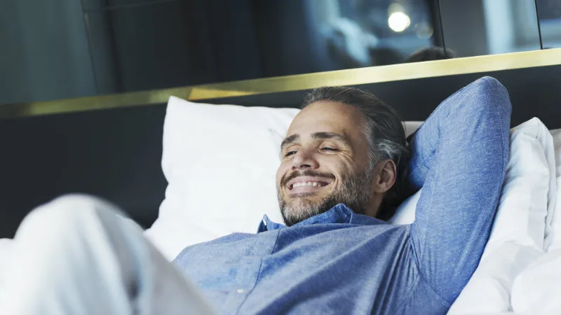 En man tar det lugnt på en hotellsäng i ett hotellrum med ett leende på läpparna.