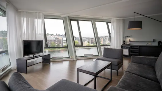 Et moderne opholdsrum med grå møbler, et TV på et stativ og store vinduer med udsigt over byens vandlandskab. Minimalistisk indretning og rigelig naturlig belysning skaber en rolig atmosfære.