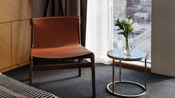 Et koselig hotellromhjørne med en brun stol, et lite bord med blomster og et stort vindu med utsikt over byen.