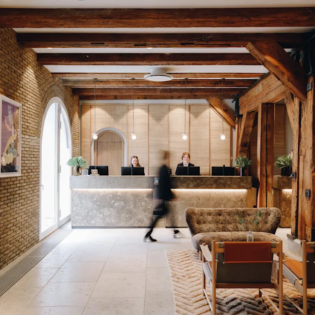 En person går gjennom en hotellobby med resepsjon, ansatte, murvegger og trebjelker, noe som skaper en varm atmosfære.
