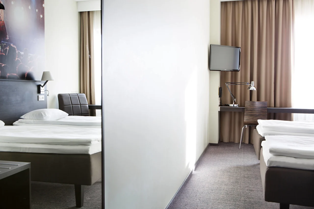 Two neatly made beds are on opposite sides of a light-filled hotel room, separated by a partial wall. A desk with a lamp, chair, and TV is visible.