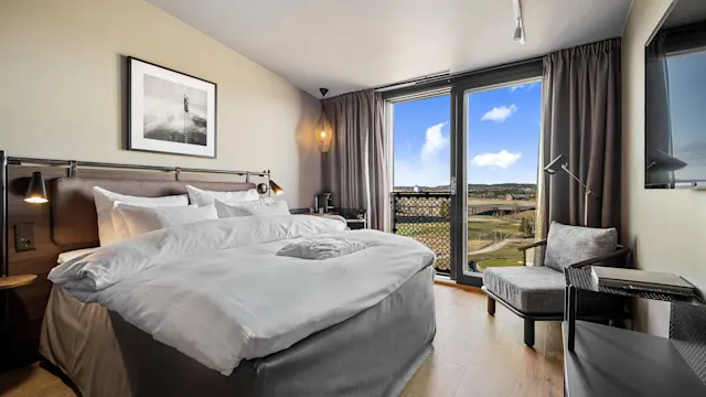 En pent bäddet seng med hvitt sengetøy er plassert på et hotellrom, mot store vinduer. Utenfor vinduene er det utsikt over et naturskjønt landskap under en delvis skyet himmel.
