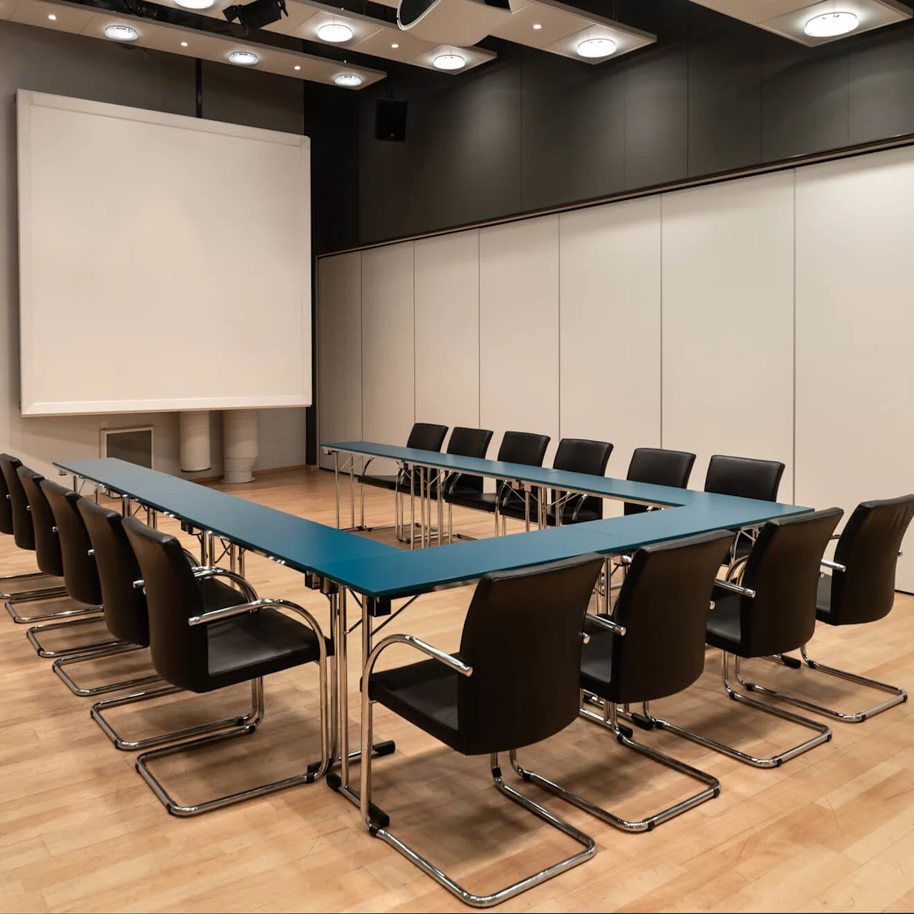 Ett U-format bord omgivet av svarta stolar står i ett modernt konferensrum med trägolv och en stor projektorduk.