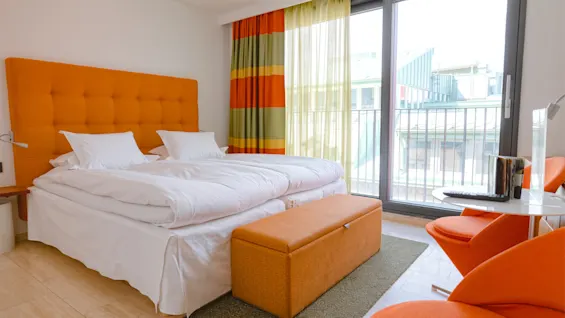 Et lyst hotellrom med en stor seng, oransje detaljer og balkongutsikt, designet for komfort og avslapning.