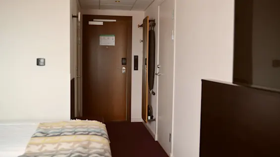 Et hotelværelse med en seng til venstre, en brun dør i midten med et skilt, og et vægmonteret strygebræt til højre.