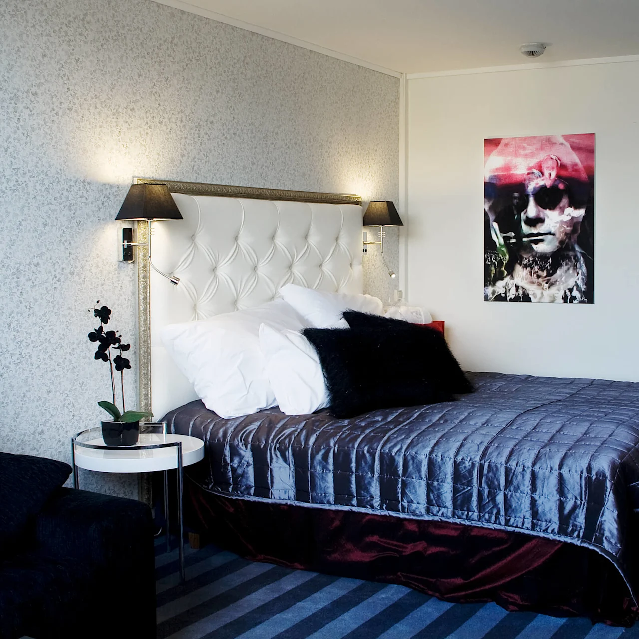 A neatly made bed with a white headboard, flanked by lamps, in a room with striped carpet and an abstract picture on the wall.