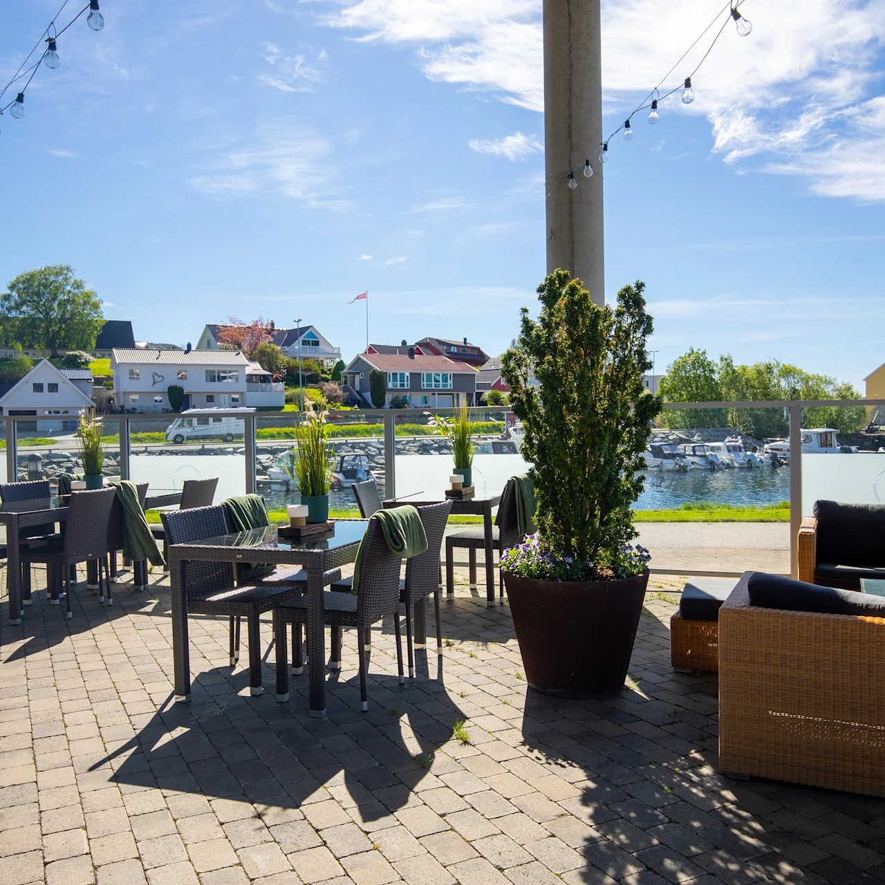 Uteplassen har kurvstoler og bord under lysslynger. Utsikt over en marina med båter og fargerike hus under en klar, solfylt himmel. Avslappet atmosfære ved vannet.