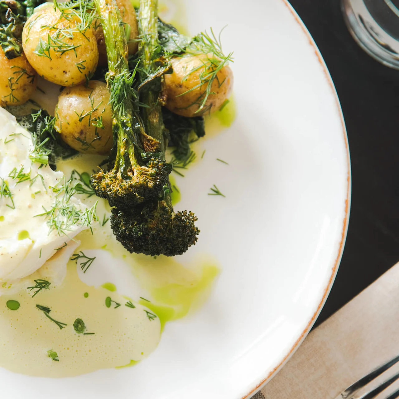 Panert hvit fisk med poteter og brokkoli toppet med fersk dill på en hvit tallerken, omgitt av en lysegrønn saus, pyntet med grønne urter.