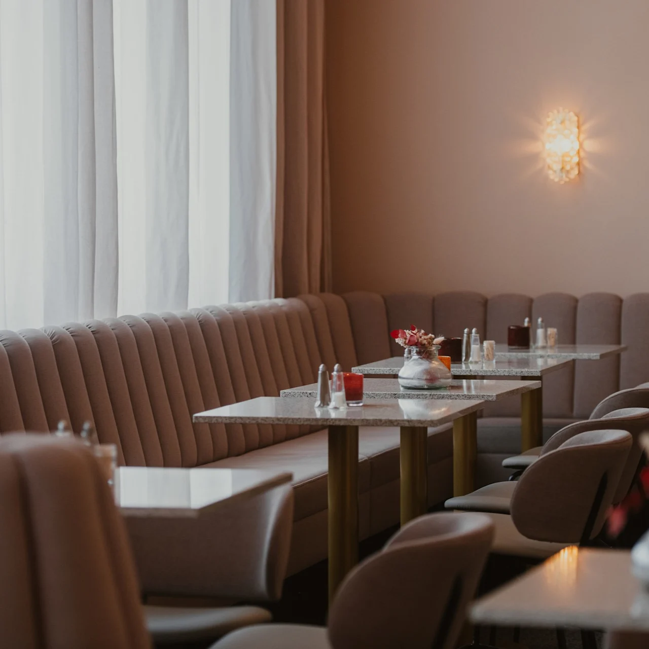 Cushioned booth seating with gray tables displaying decorative vases and condiments. Soft lighting and tall curtains create a cozy cafe ambiance.