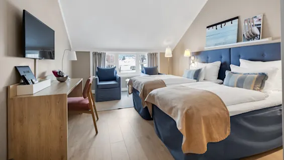 Et hotellrom med to senger dekket med beige tepper og blå hodegjerder, flankert av lamper. Et skrivebord med en velkomstbrett vender mot et vindu med snølandskap.