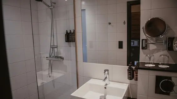 Moderni hotellihuoneen kylpyhuone, jossa on lasitettu suihku, neliönmuotoinen pesuallas ja suuri peili, käyttövalmiina.