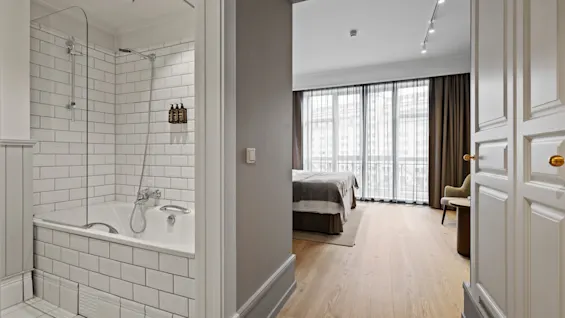 A modern hotel room with a bathroom featuring a bathtub and a bedroom with a large bed and bright windows.