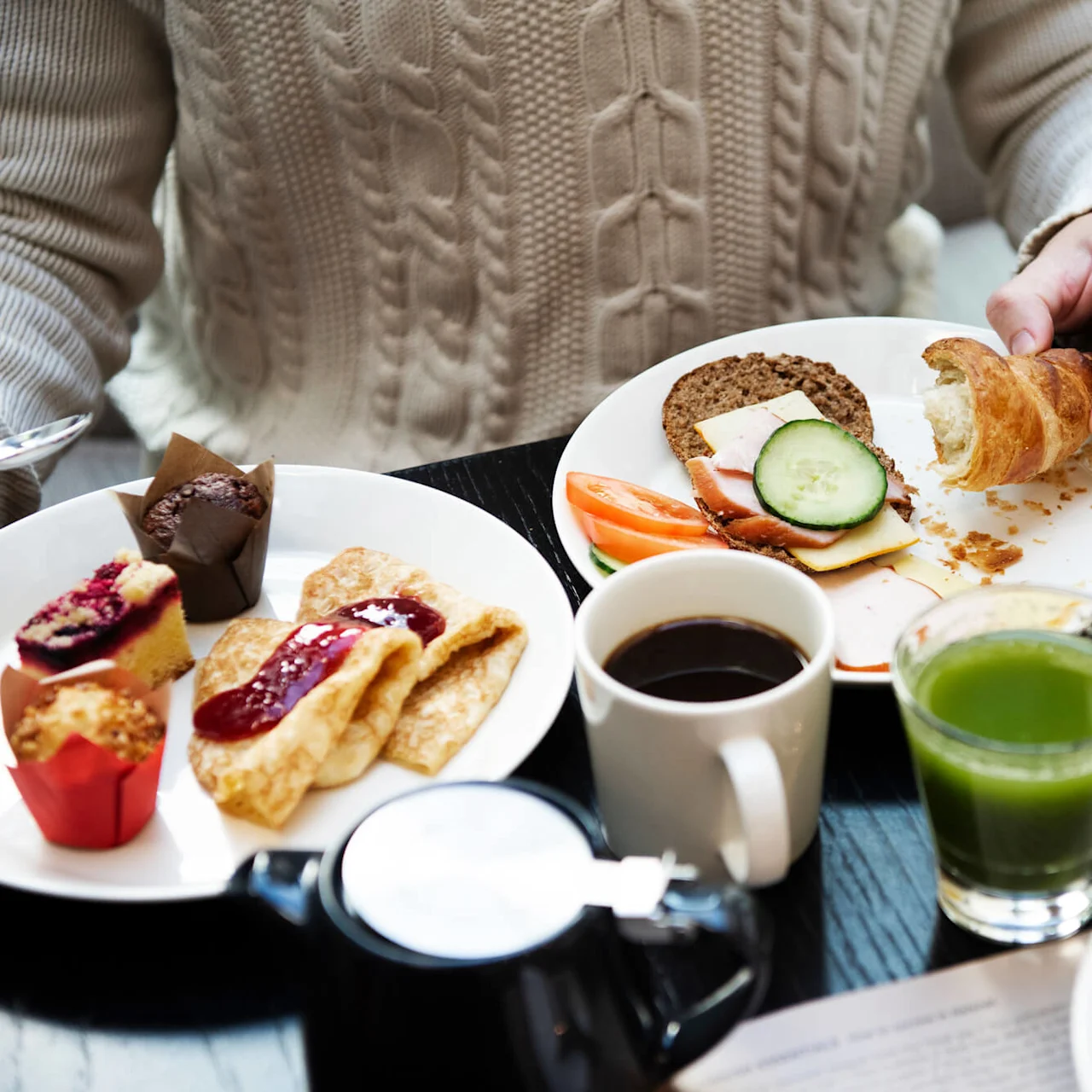 En person i en genser nyter en frokost med bakverk, smørbrød, frukt, kaffe og juice på et mørkt bord.
