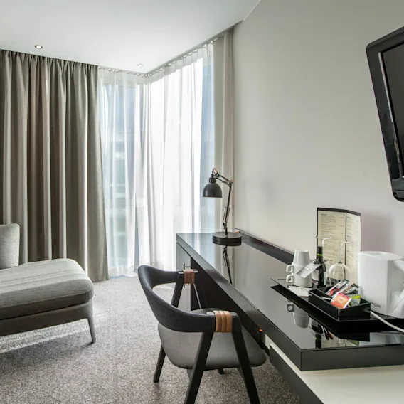 A modern hotel room features a gray chaise lounge with pillows, a desk with a lamp and tray, and floor-to-ceiling curtains filtering sunlight.