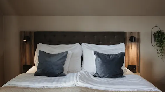 Viihtyisä hotellisänky valkoisilla liinavaatteilla, harmailla tyynyillä ja lämpimällä valaistuksella, valmiina rauhalliseen yöhön.