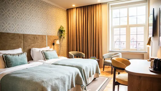 Två prydligt bäddade sängar med ljusblå filtar är placerade bredvid ett fönster med fördragna gardiner i ett varmt upplyst hotellrum. Ett skrivbord och stolar kompletterar möbleringen.