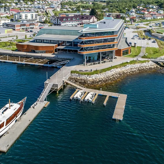 Et moderne hotell ved sjøen med en privat brygge og båter under en klar blå himmel.