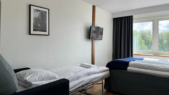 A sofa bed with pillows and a blanket is unfolded beside a double bed, in a simple room featuring a wall-mounted TV, framed artwork, and large window with garden view.