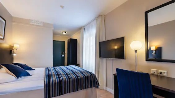 A neatly arranged hotel room features a double bed with blue-striped bedding, a wall-mounted TV, and a desk with a lamp. Light wood flooring complements beige walls.