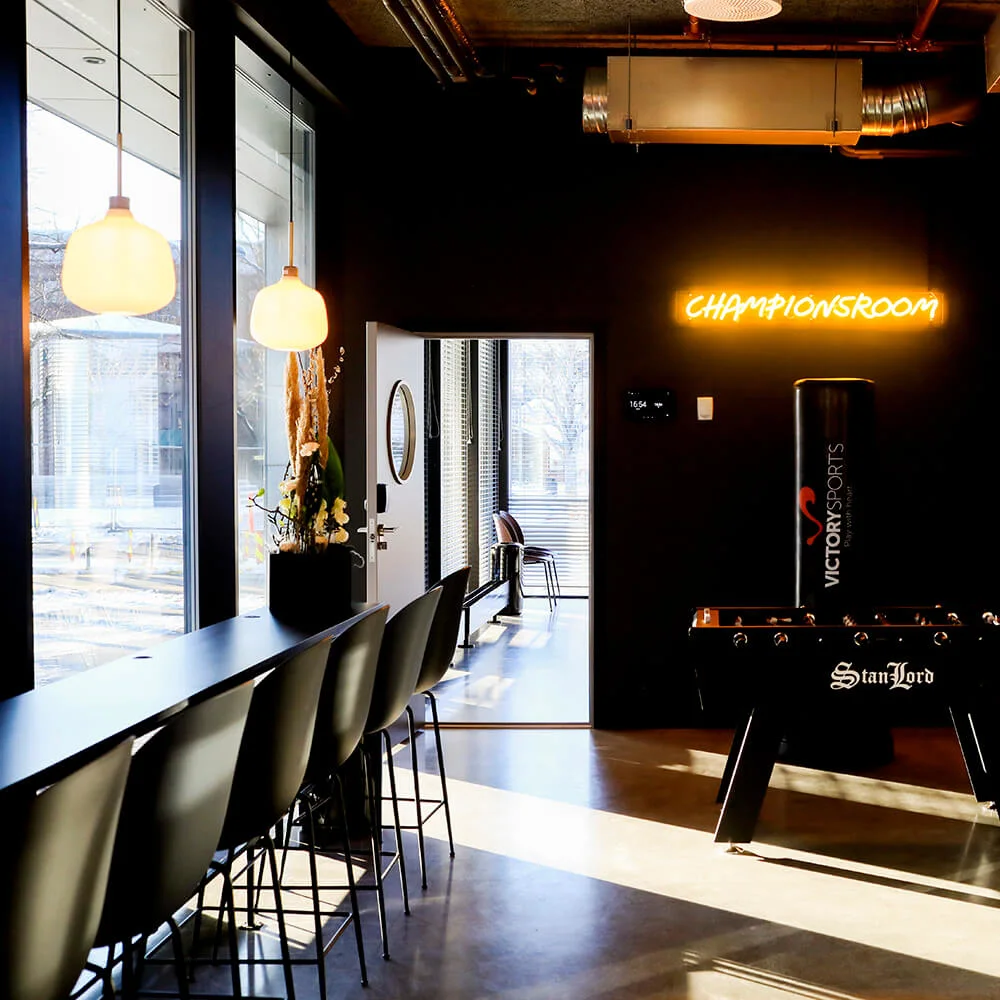 Bordfodbold står i et moderne rum med mørke vægge og et neonskilt, der siger "CHAMPIONSROOM." Store vinduer lukker naturligt lys ind og oplyser barstole og dekorative planter.