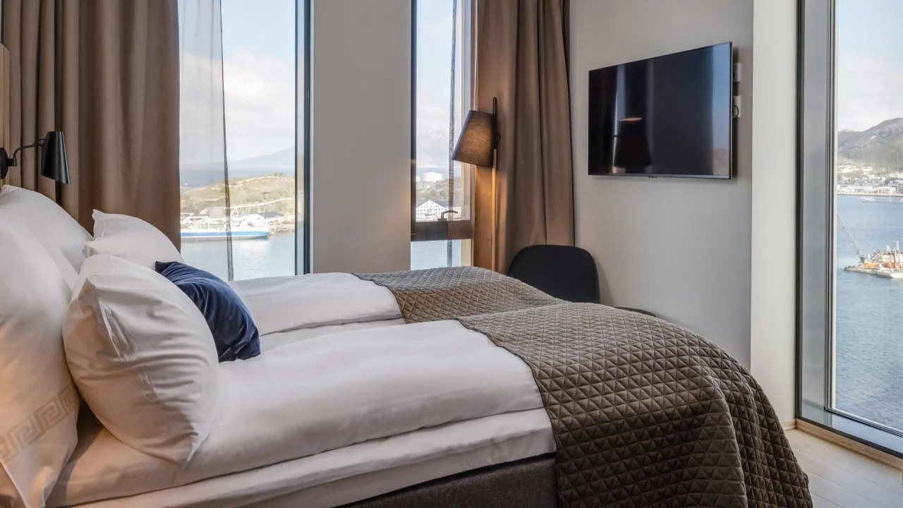 A modern bedroom with a large window offering a view of a harbor with boats, under a clear sky.