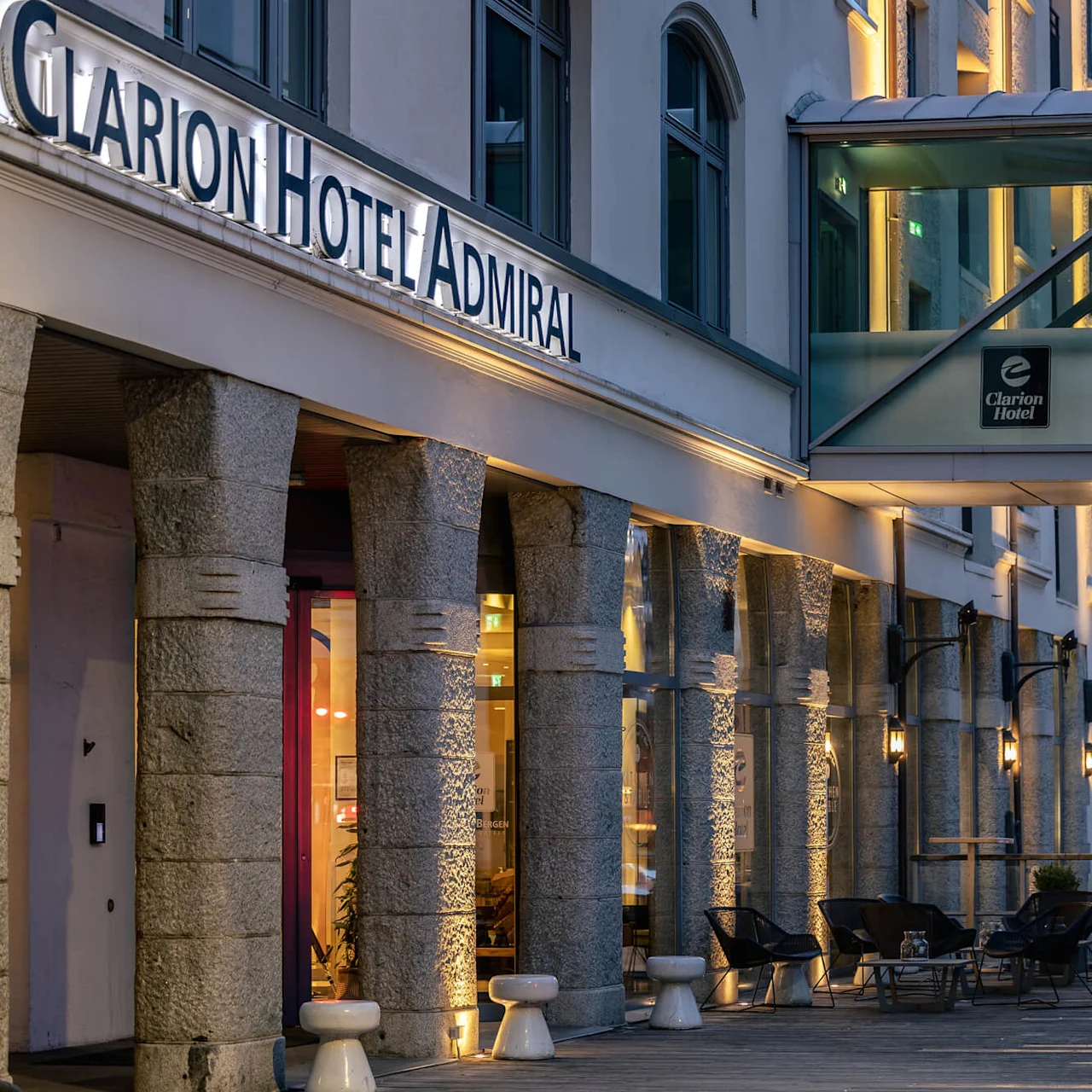 Indgangen til Clarion Hotel Admiral under en overdækket gangbro, med et skilt der står "SMALL TALK". Siddepladser og udendørsbelysning skaber en indbydende aftenstemning.