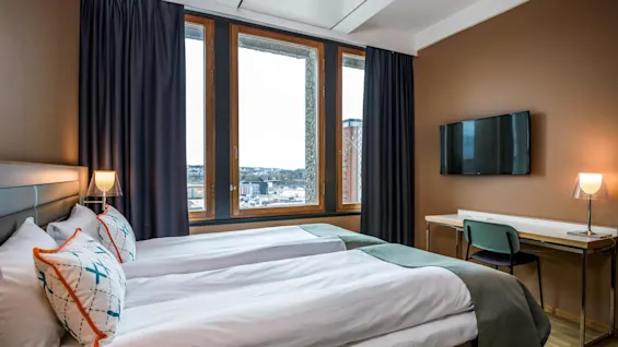 A neatly made bed with patterned pillows sits near a window with curtains, overlooking a cityscape. A desk with a chair and lamp faces a wall-mounted TV.