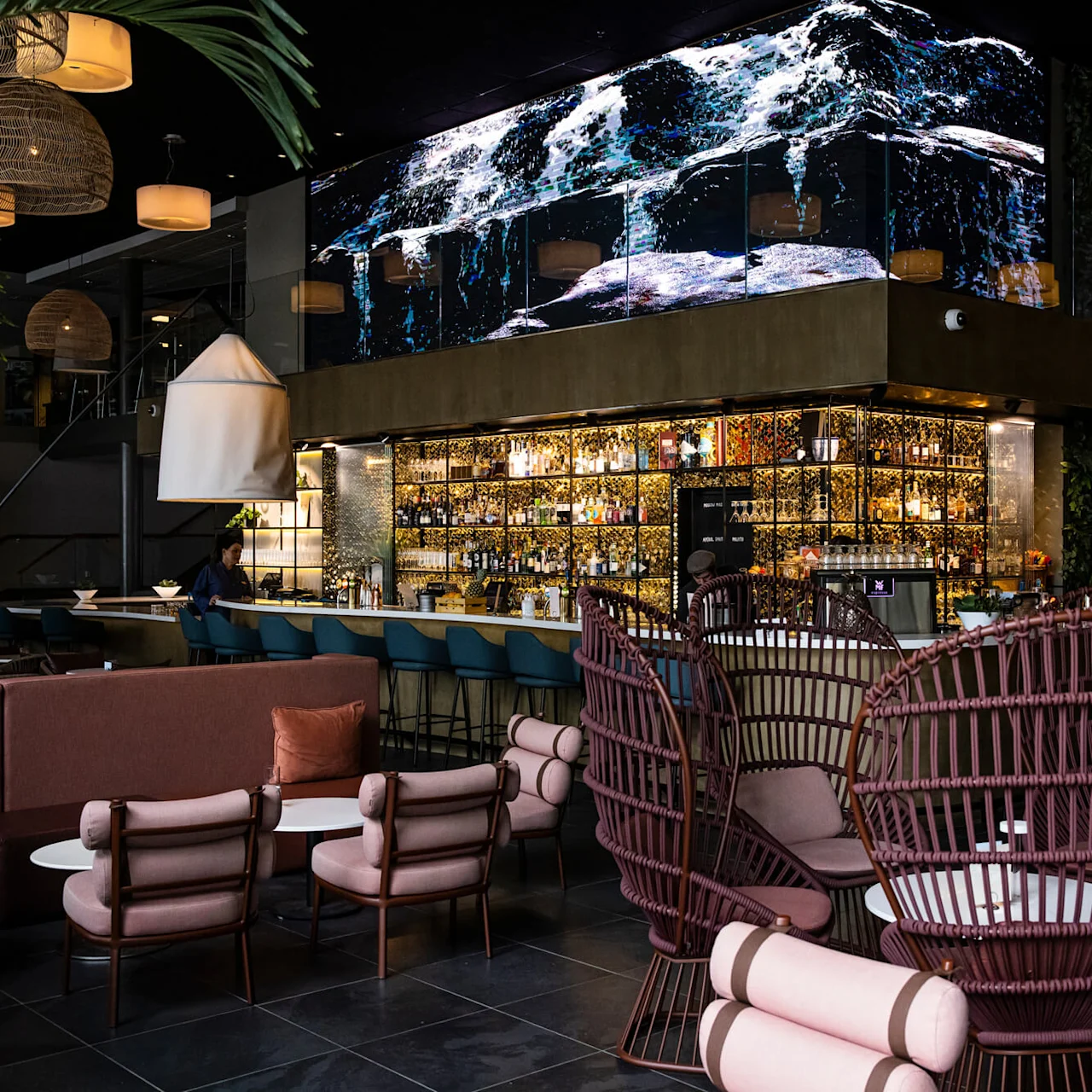 En moderne bar med en stor skjerm som viser bølger over et fylt bar, omgitt av eklektiske sitteplasser og hengelamper, som skaper en stilfull, sofistikert atmosfære.