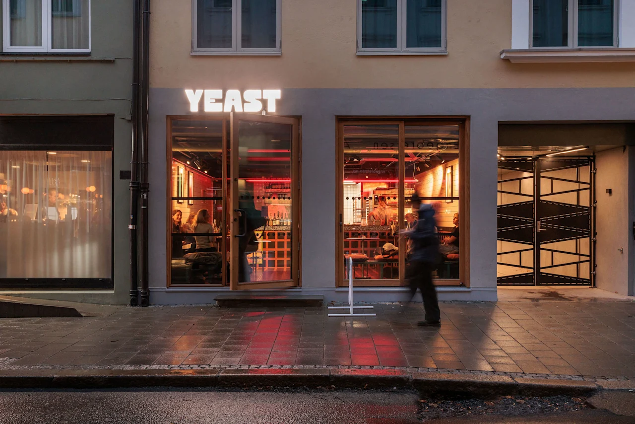 A vibrant restaurant Yeast at dusk with people dining inside, and a person walking on the wet sidewalk outside.