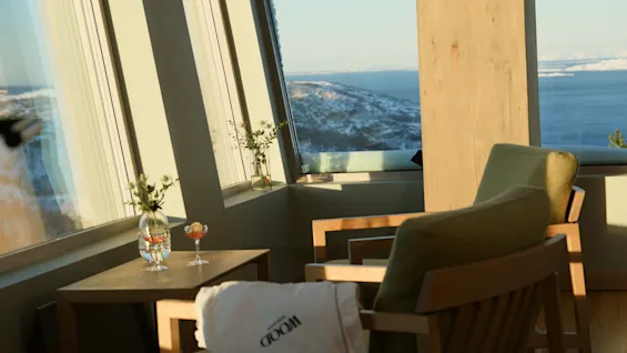 Et hyggeligt siddeområde med store vinduer, der vender ud mod et snedækket landskab og vand. To stole, et bord med drikke og blomster.