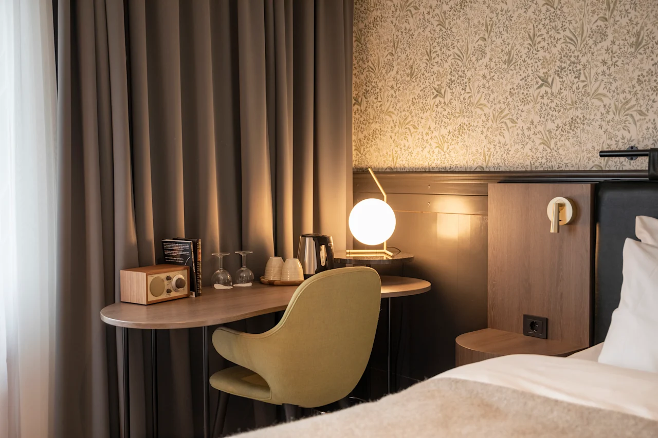 Et skrivebord med en grønn stol har en radio, glass og en sfærisk lampe, ved siden av en seng. Mørke gardiner og mønstret tapet skaper en koselig romatmosfære.