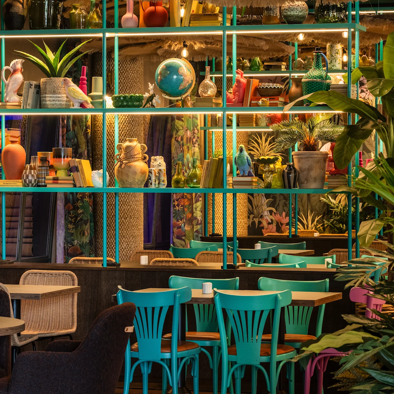 Shelves displaying colorful vases, plants, and a globe in a vibrant cafe setting with wicker chairs and teal tables, surrounded by lush greenery.