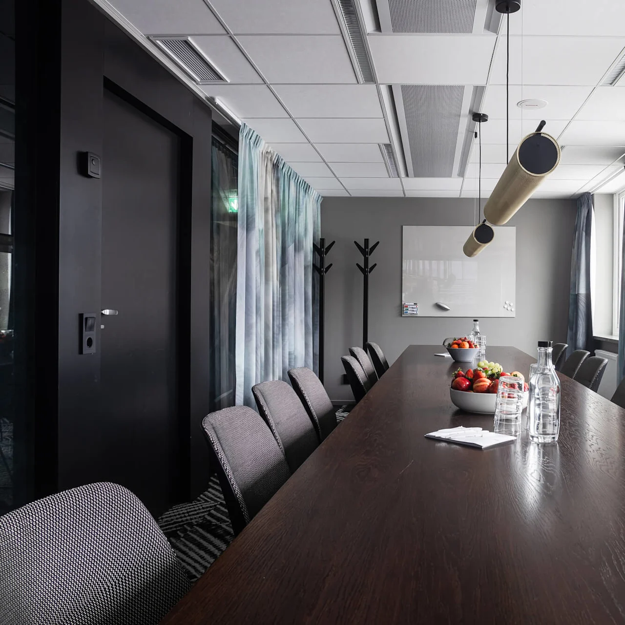 Ett långt konferensbord med stolar, med tallrikar med frukt och vattenflaskor, är omgivet av gardiner, klädhängare och stora fönster i ett modernt mötesrum.