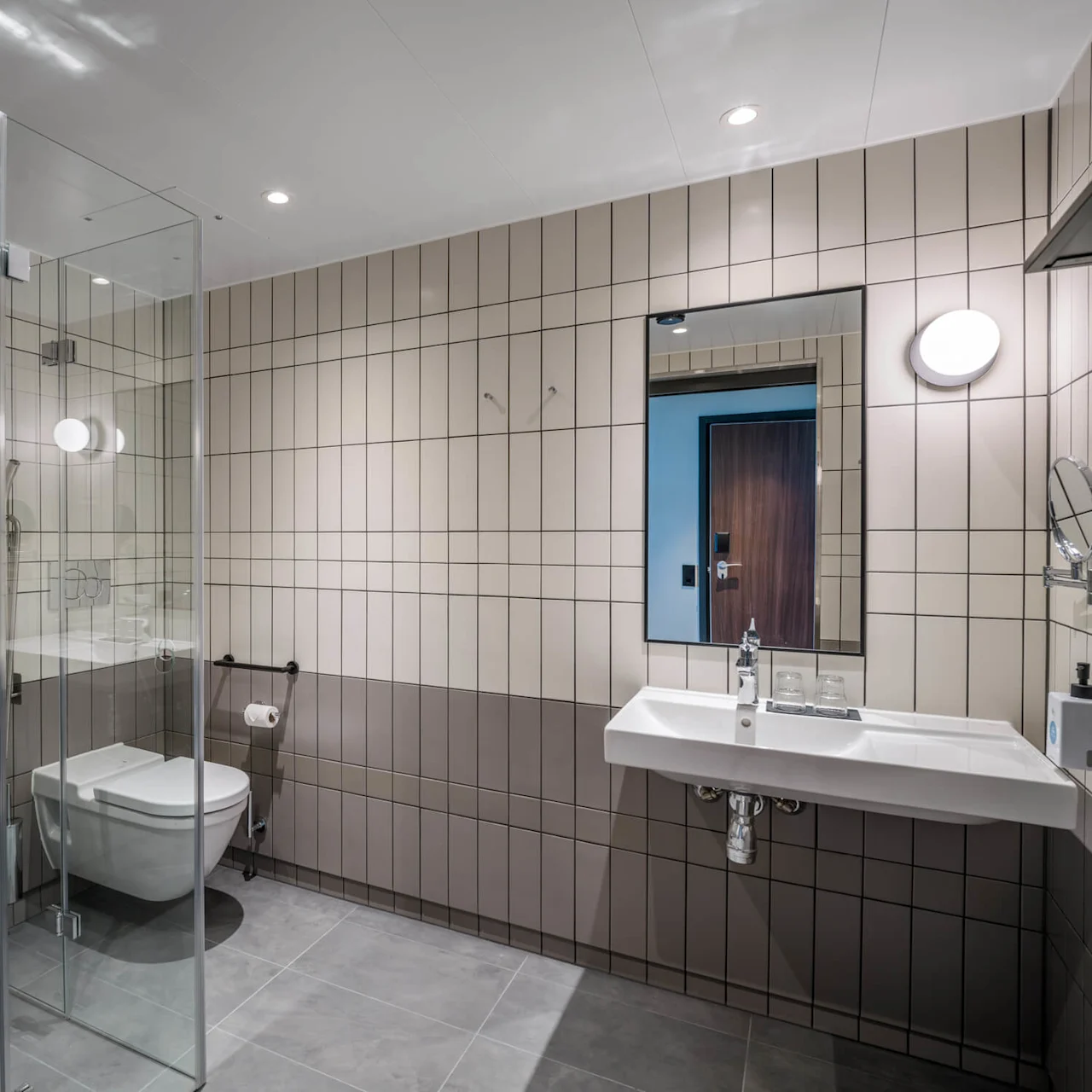 Et moderne bad har en glassinnrammet dusj, veggmontert toalett og bredt servant under et speil. Beige fliser dekker veggene, og skaper et rent og moderne rom.