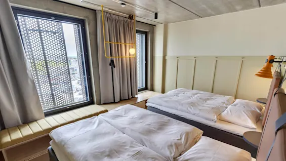 To pent reddede senger med hvitt sengetøy står i et moderne rom med store vinduer dekket av perforerte paneler som gir utsikt over byen. Grå gardiner og en gul lampe tilfører dekor.
