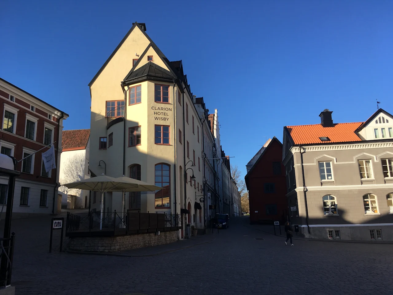 Clarion Hotel Wisby, med sin beige facade og høje buede vinduer, står på en brostensbelagt gade under en klar blå himmel. Nærliggende bygninger kaster skygger på jorden.