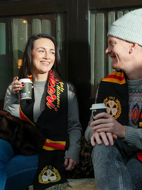 Två leende sportfans som sitter utomhus i vinterkläder och njuter av varm dryck tillsammans.