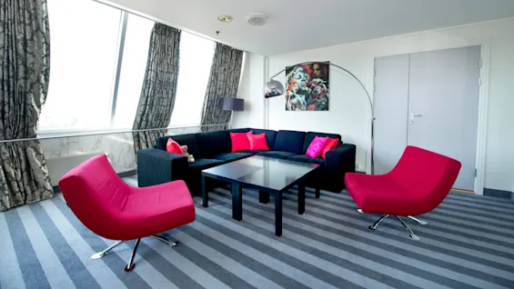 A modern living room with a large blue sofa, red accent chairs, and striped carpet, bathed in natural light from large windows.