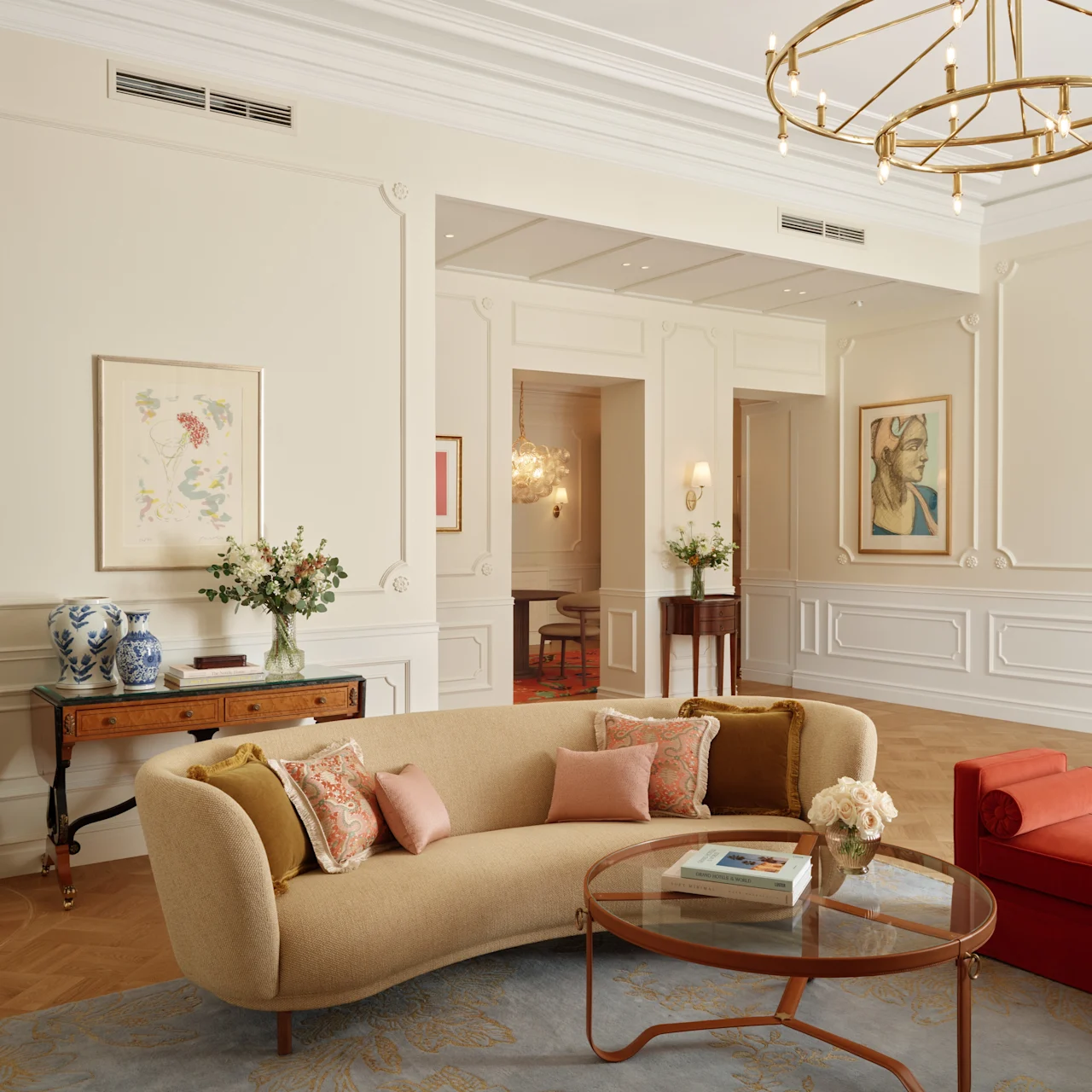 A beige curved sofa sits with decorative pillows and a glass coffee table at the center of an elegant, well-lit room, featuring art, a red chaise, and a grand piano.