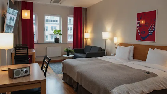 A neatly made double bed with pillows and beige blanket in a modern hotel room. Lamps illuminate the space, adorned with red curtains, a small sofa, and a pop art wall poster.