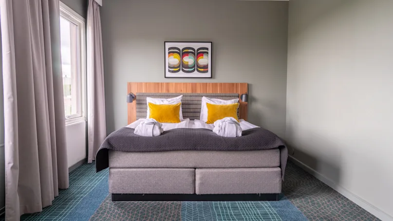 A neatly made double bed with two yellow cushions and folded white towels rests against a headboard. Three abstract paintings hang above; sheer curtains frame a window on the left.