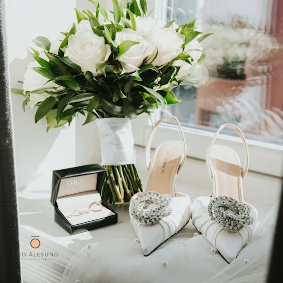 Bryllupsdetaljer i en vinduskarm med en bukett hvite roser, elegante hvite brudesko med krystallspenner og en eske med gifteringer.