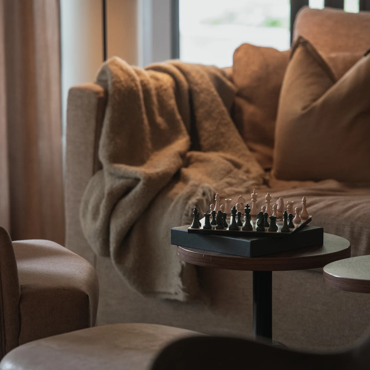 A chess set rests on a small round table. Surrounding it are a cozy sofa with cushions and a draped blanket, creating a warm, inviting living room ambiance.