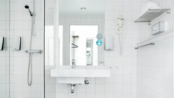 Ett vitt kaklat badrum har ett väggmonterat handfat med spegel ovanför, bredvid en glasdusch, hårtork, handdukshängare och servetthållare. En skylt lyder "USE LESS STUFF" och "THINK MORE WATER."