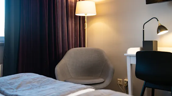 A gray chair sits under a lit floor lamp beside dark curtains. Nearby, a desk holds a black lamp and chair, in a softly illuminated room.