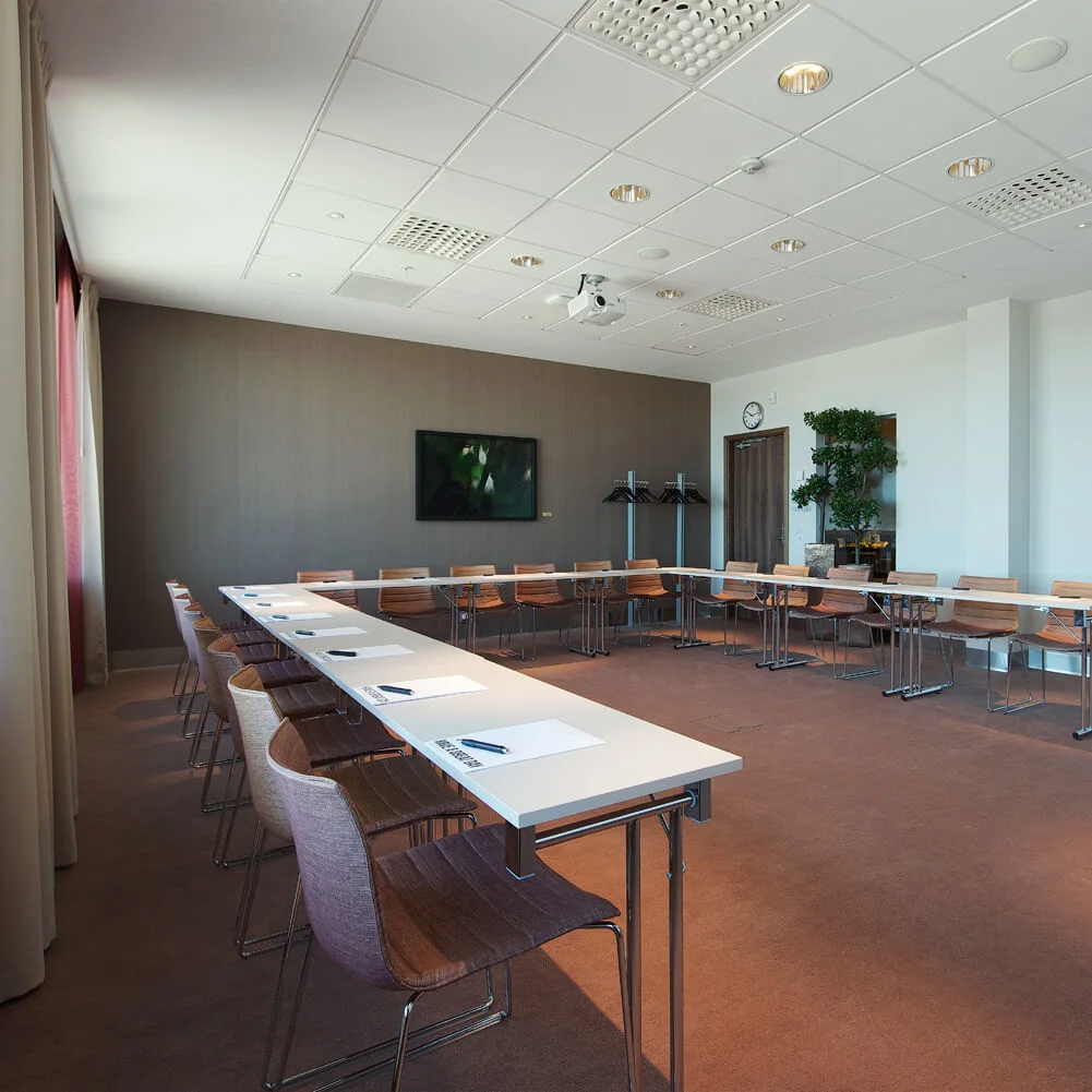 Meeting tables are arranged in a U-shape with chairs, papers, and pens placed on them. A wall-mounted TV, clock, and coat rack are in the background.